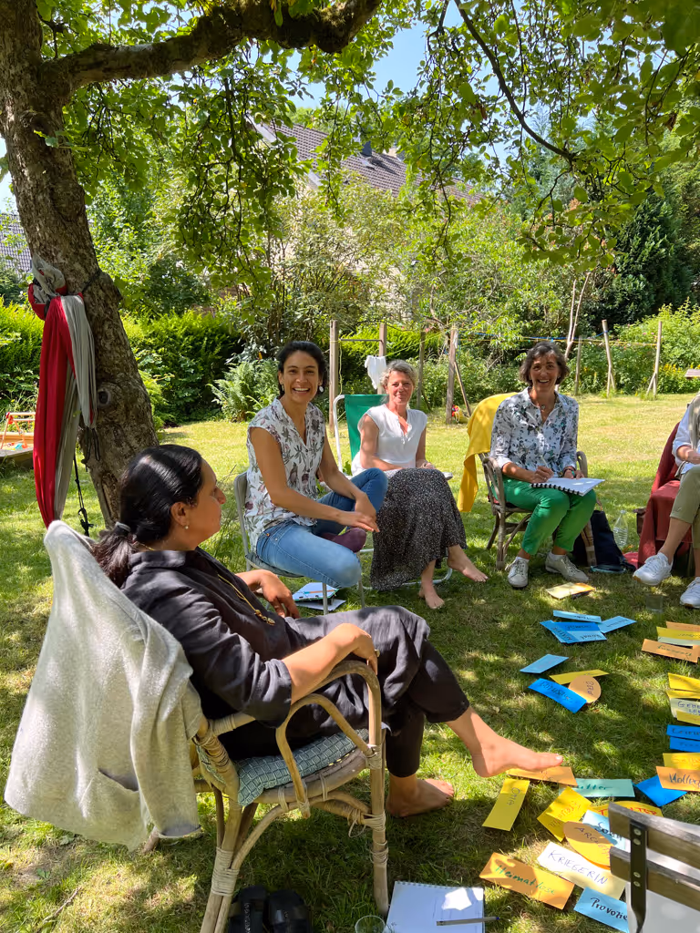 Group of women sitting in a circle outdoors on grass under a tree, smiling and engaged in discussion with colorful paper cards spread on the ground.